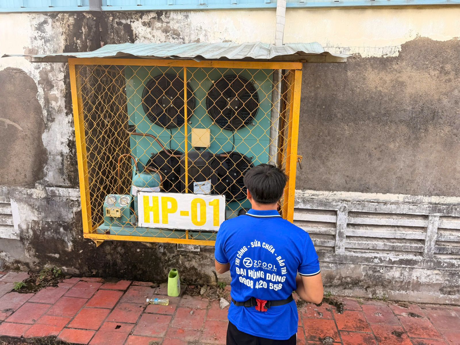 Sửa Máy Lạnh Công Nghiệp: Cụm máy mang biển HP-01 trong lồng bảo vệ và nhân sự đứng trước thiết bị.