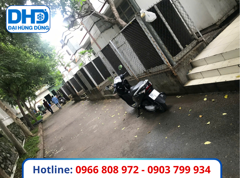 Thanh Lý Hệ Thống Máy Lạnh Công Nghiệp: Dãy thiết bị ngoài nhà phía sau hàng rào lưới, chạy dọc lối đi có cây xanh.